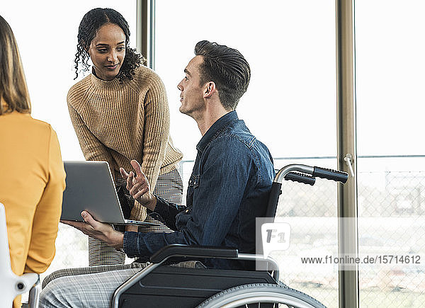 Young businessman in wheelchair showing laptop to colleague in office