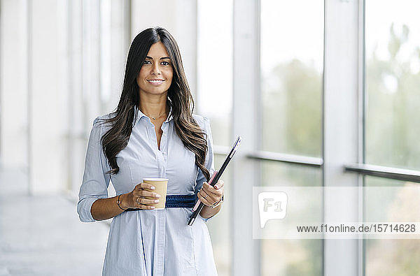 Porträt einer lächelnden Geschäftsfrau mit Tablette und Kaffee zum Mitnehmen am Fenster