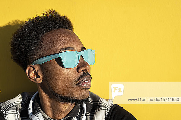 Porträt eines jungen Mannes mit hellblauer Sonnenbrille vor gelber Wand