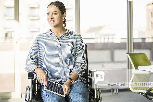 Young businesswoman with tablet sitting in wheelchair in office