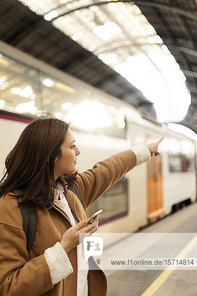 Junge Frau mit Handy auf dem Bahnhof  die mit dem Finger zeigt