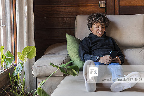 Lächelnder entspannter Junge  der sein Handy zu Hause auf der Couch benutzt