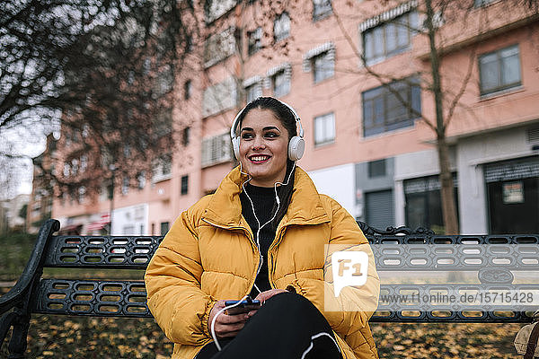 Porträt einer glücklichen jungen Frau  die im Herbst auf einer Bank sitzt und mit Kopfhörer und Smartphone Musik hört