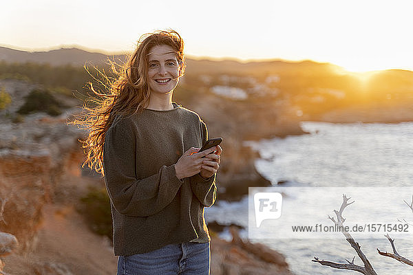 Porträt einer lächelnden rothaarigen jungen Frau mit Handy an der Küste bei Sonnenuntergang  Ibiza  Spanien
