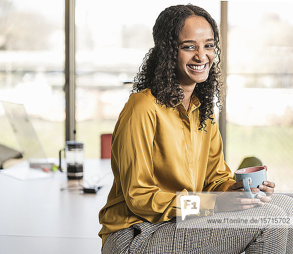 Porträt einer glücklichen jungen Geschäftsfrau  die im Büro am Schreibtisch sitzt und eine Kaffeepause macht