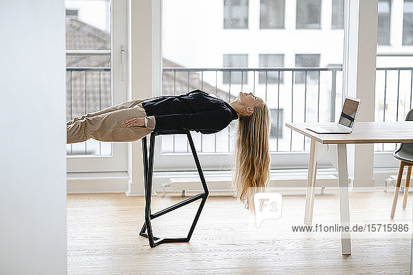 Junge Geschäftsfrau  die im Büro auf einem Hocker liegend Yoga praktiziert