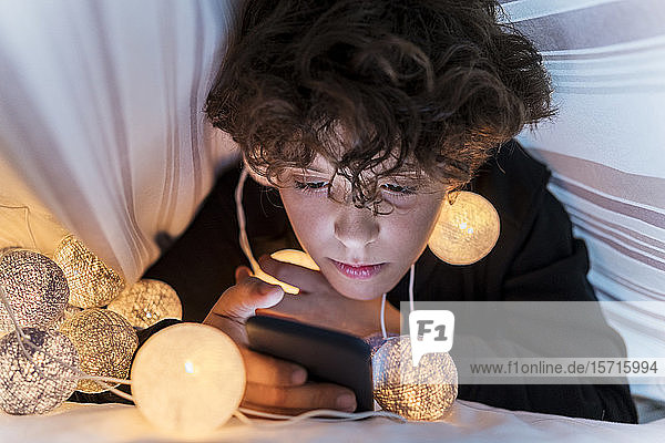 Junge mit Lichterkette unter der Bettdecke  der sein Handy benutzt