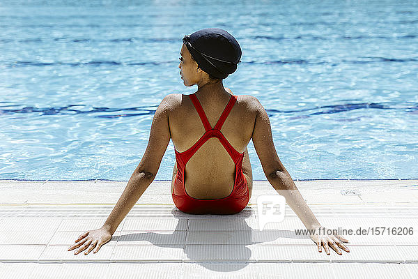 Rückenansicht einer jungen Frau in rotem Badeanzug und Badekappe  die sich nach dem Schwimmen am Pool entspannt