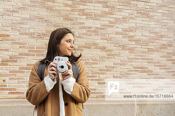 Junge Frau mit Kamera vor einer Ziegelmauer  die sich umsieht