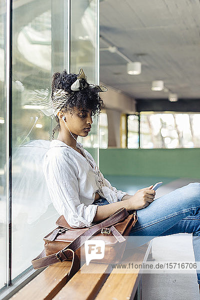 Porträt einer jungen Frau mit Ohrstöpseln  die auf einer Bank sitzt und auf ein Mobiltelefon schaut