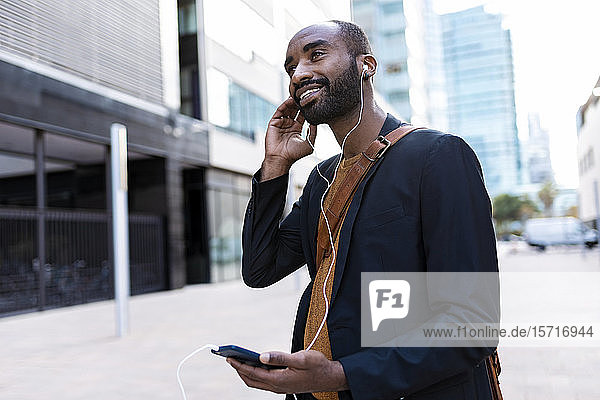 Lächelnder junger Geschäftsmann  der in der Stadt mit Kopfhörern und Smartphones Musik hört