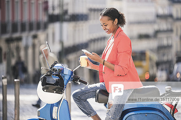 Smiling young woman using cell phone on motor scooter in the city  Lisbon  Portugal