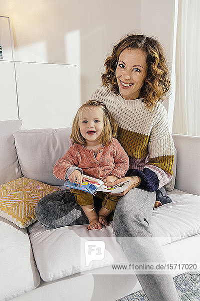Porträt einer glücklichen Mutter und einer kleinen Tochter mit Buch auf der Couch zu Hause
