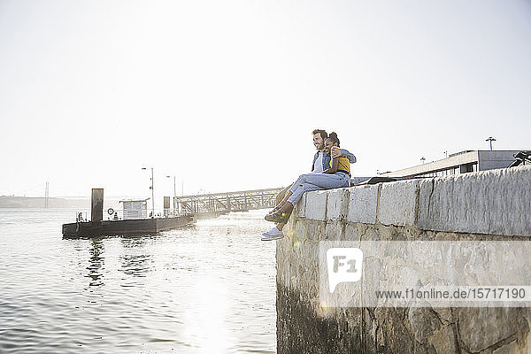 Young couple sitting on pier at the waterfront enjoying the view  Lisbon  Portugal