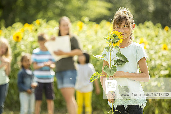 Kleines Mädchen hält Sonnenblume  im Hintergrund stehen Lehrer und Freunde