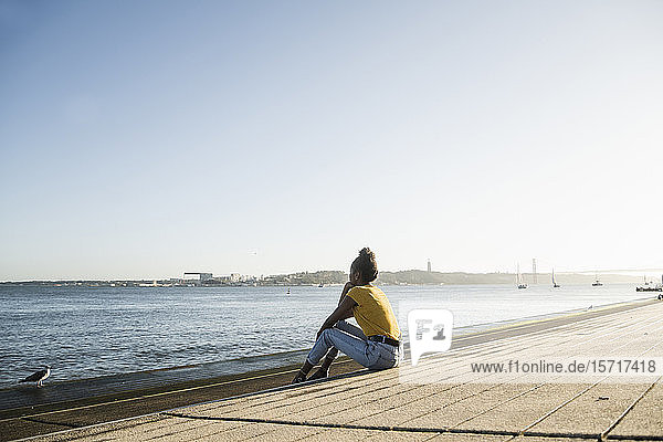 Rear view of young woman sitting at the waterfront at sunset  Lisbon  Portugal