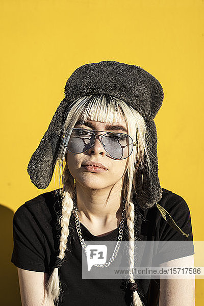 Porträt einer jungen Frau mit gefärbten Haaren  die eine Mütze und eine Sonnenbrille gegen eine gelbe Wand trägt