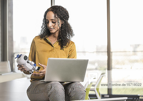 Junge Geschäftsfrau sitzt im Büro mit Laptop und Roboter auf dem Schreibtisch