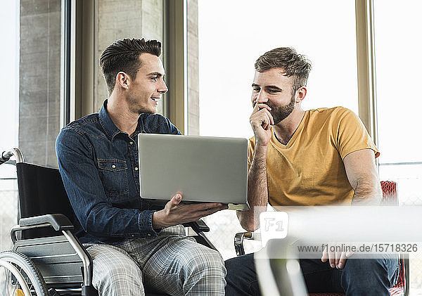 Young businessman in wheelchair showing laptop to colleague in office