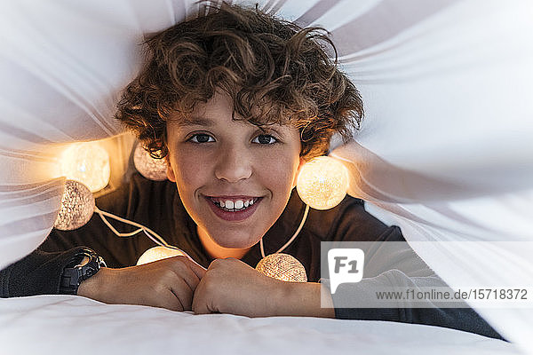 Porträt eines glücklichen Jungen mit Lichterkette unter der Bettdecke