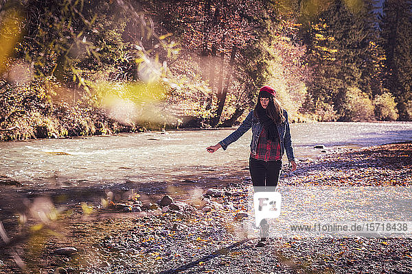 Frau mit rotem Wollhut und Jeansjacke geht im Herbst am Flussufer spazieren