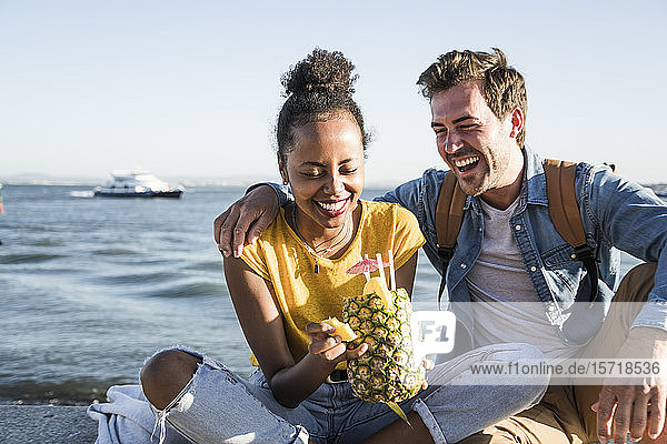 Happy young couple sitting on pier at the waterfront with a pineapple  Lisbon  Portugal