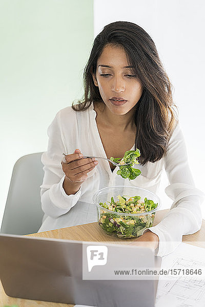 Porträt eines jungen Architekten  der am Schreibtisch gemischten Salat isst und auf seinen Laptop schaut