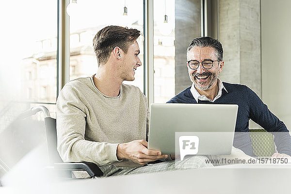 Happy mature businessman and young man in wheelchair using laptop in office