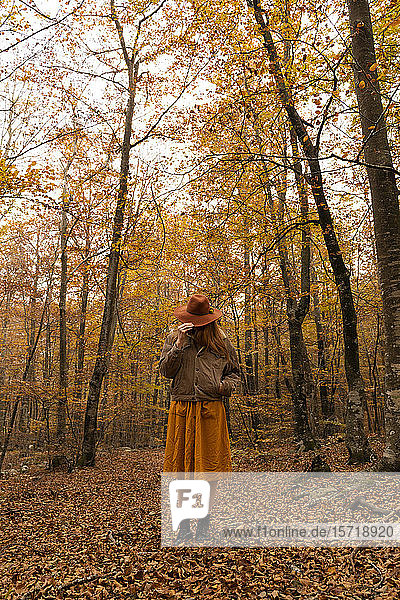 Modische rothaarige junge Frau im herbstlichen Wald  die ihr Gesicht mit einem Hut bedeckt