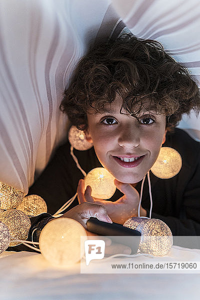 Porträt eines lächelnden Jungen mit Lichterkette und Handy unter der Bettdecke