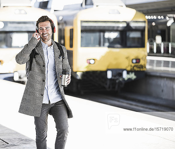 Glücklicher junger Geschäftsmann mit Kaffeetasse und Kopfhörern geht am Bahnhof spazieren