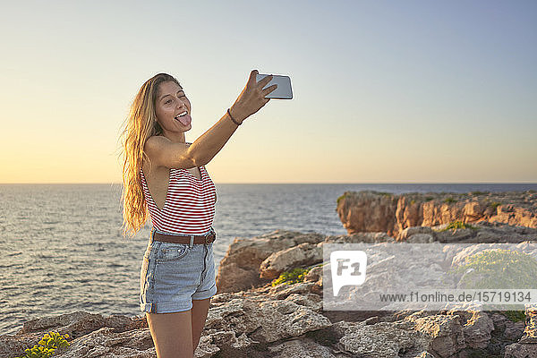 Junge Frau steht auf einer Klippe am Meer  benutzt ein Smartphone  nimmt sich selbst
