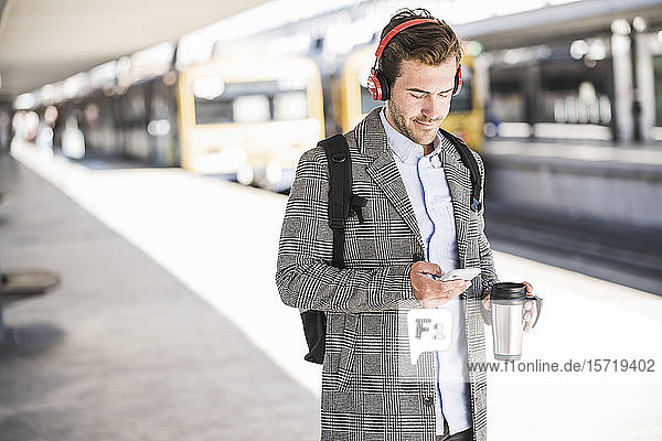 Junger Geschäftsmann mit Handy und Kopfhörern am Bahnhof