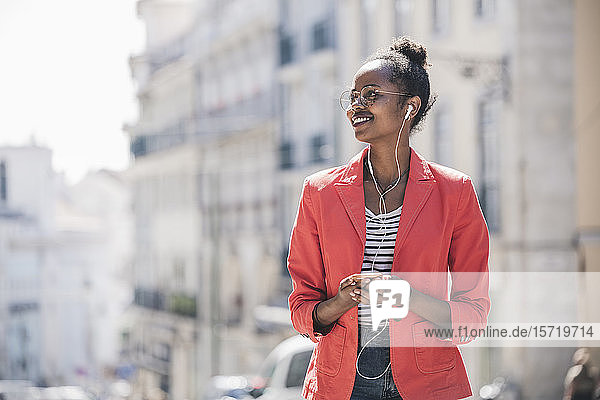 Lächelnde junge Frau mit Kopfhörern und Smartphone  die sich in der Stadt umsieht  Lissabon  Portugal