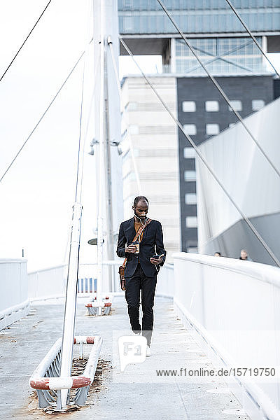 Junger Geschäftsmann mit Kopfhörern und Kaffee zum Spaziergang auf dem Steg mit Blick auf Smartphone