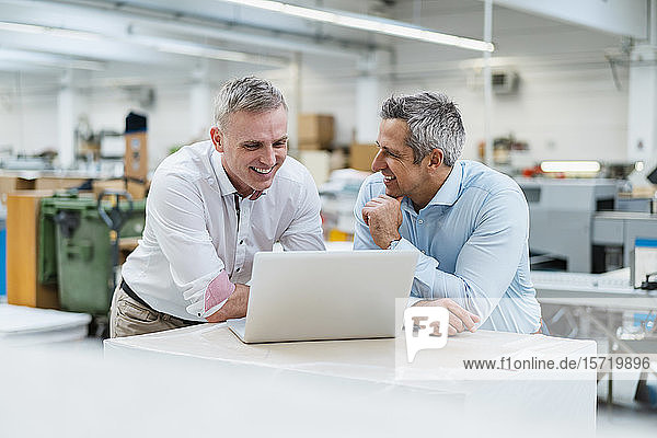 Zwei lächelnde Kollegen am Laptop und diskutieren in einer Fabrik