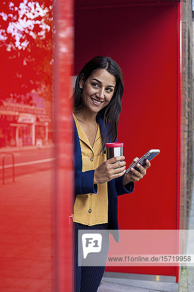 Junge Geschäftsfrau mit wiederverwendbarer Kaffeetasse und Smartphone   an die Wand gelehnt