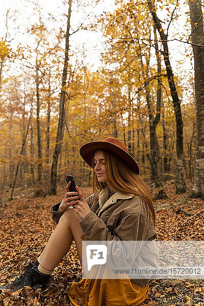 Fashionable redheaded young woman sitting in autumnal forest looking at cell phone