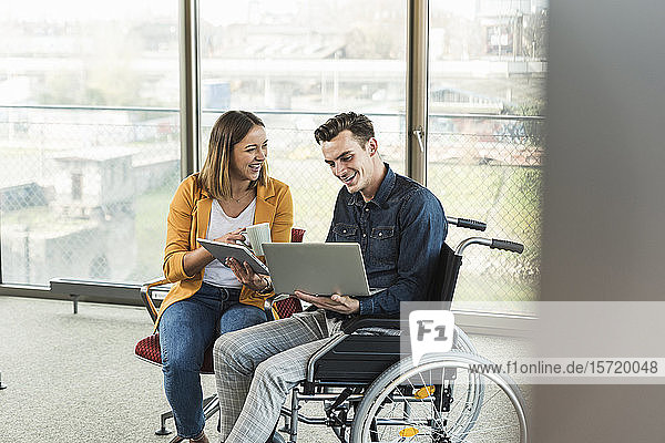 Happy young businessman with laptop in wheelchair and businesswoman with tablet in office