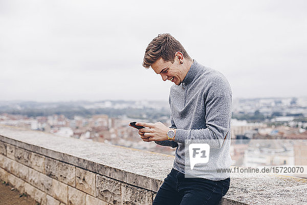 Blonder junger Mann mit einem Pullover  der in Lleida lächelnd eine Nachricht auf sein Handy schreibt