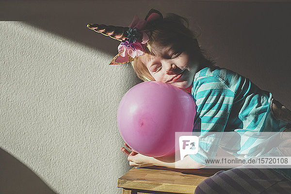 Porträt eines kleinen Mädchens mit geschlossenen Augen  das ein Einhorn-Horn trägt und sich auf einen rosa Ballon stützt