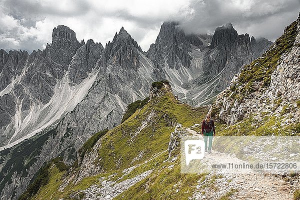 Junge Frau  Bergsteigerin auf einem Wanderweg  hinter Bergspitzen und scharfen Felsgipfeln  dramatische Wolken  Cimon di Croda Liscia und Cadini-Gruppe  Sextner Dolomiten  Belluno  Italien  Europa
