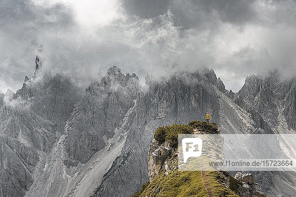 Frau in gelber Jacke auf einem Bergrücken stehend  hinter ihr Bergspitzen und scharfe Felsgipfel  dramatische Wolken  Cimon die Croda Liscia und Cadini Gruppe  Auronzo di Cadore  Belluno  Italien  Europa