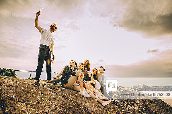 Man taking selfie with friends on rock formation against sky in picnic during sunset