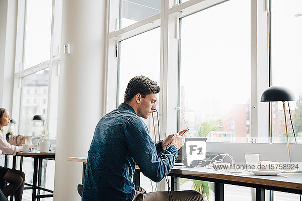 Junger Geschäftsmann nutzt Smartphone  während er mit Laptop am Schreibtisch im Büro sitzt