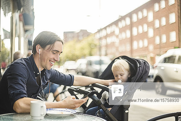 Lächelnder Mann hört Musik über Kopfhörer von einem Smartphone  während er mit seinem Baby im Straßencafé in der Stadt sitzt