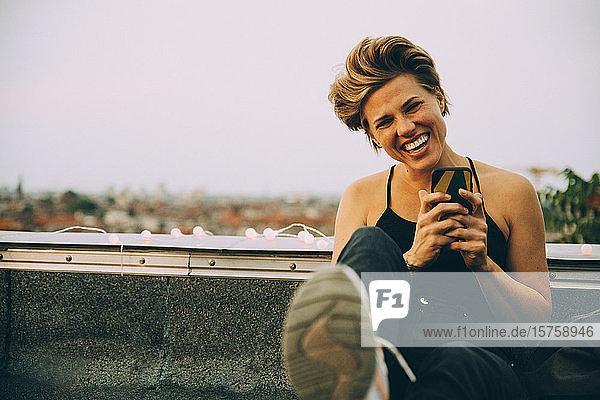 Porträt einer fröhlichen Frau  die ein Mobiltelefon benutzt  während sie auf einer Terrasse gegen den Himmel sitzt