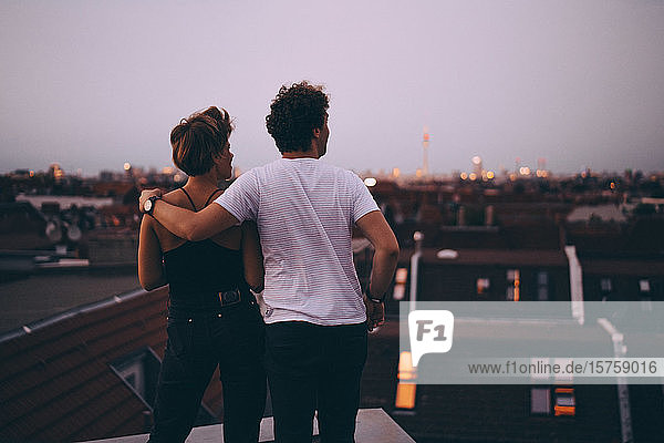 Rückansicht eines Paares  das mit dem Arm um die Stadt blickt  während es in der Dämmerung auf der Terrasse steht