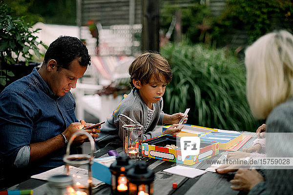 Familie spielt Brettspiel am Tisch  während der Vater sein Handy benutzt