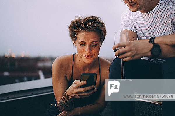 Frau benutzt Smartphone  während sie bei einem Freund sitzt und Wein auf der Terrasse trinkt